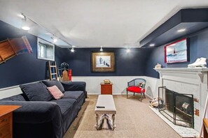 A room with a dark blue wall, a black sofa, a red chair, a white fireplace, and a framed picture on the wall. - Unsalted Tides-Walk to Beach + Town + Dog-Friendly (South Haven)