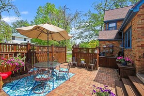 A patio with a table and chairs, a hanging flower basket, and a brick wall. - Unsalted Tides-Walk to Beach + Town + Dog-Friendly (South Haven)