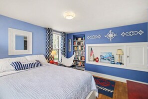 A bedroom with a bed, a bookshelf, a window with patterned curtains, and a wall-mounted shelf with decorative items. - Unsalted Tides-Walk to Beach + Town + Dog-Friendly (South Haven)