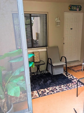 A room with a sliding glass door, a window with blinds, a small table with a chair, a welcome mat, and a door with a number plate. - Forbidden Safari - Your Dockside Paradise Lakes Stay (Lutz)