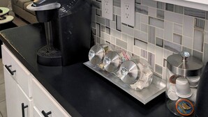 Uma cozinha com armários brancos, uma cafeteira preta e uma bandeja com copos de vidro transparentes.
