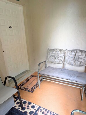 Una habitación con una puerta blanca, un banco de metal con cojín, una alfombra estampada y una mesita.