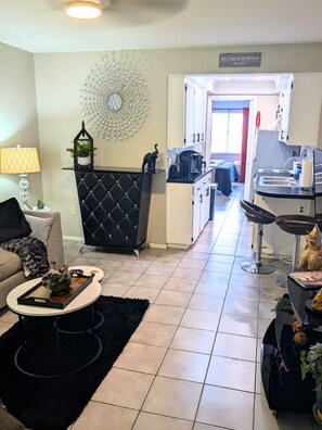 A modern living room with a black sofa, a coffee table, and a decorative mirror. A kitchen area is visible in the background with a dining table and chairs. - Forbidden Safari - Your Dockside Paradise Lakes Stay (Lutz)