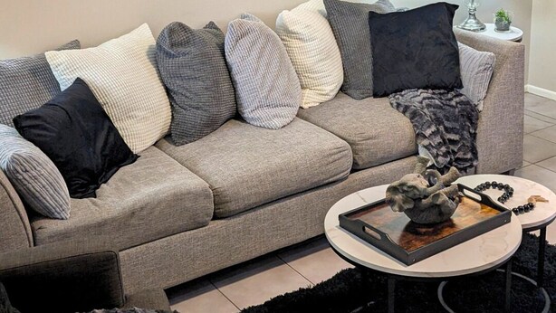 Uma sala de estar com um sofá cinza, vários almofadas, um tapete preto, uma mesa de centro com objetos decorativos, um abajur e uma obra de arte com padrão de zebra na parede.