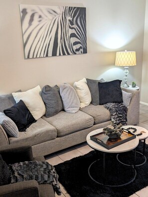 Ett vardagsrum med en grå soffa, flera kuddar, en svart matta, ett soffbord med dekorativa föremål, en lampa och ett konstverk med zebrabråmönster på väggen.