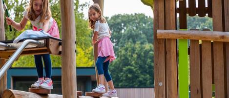 Twee kinderen spelen op een houten speeltoestel met een glijbaan en klimelementen.