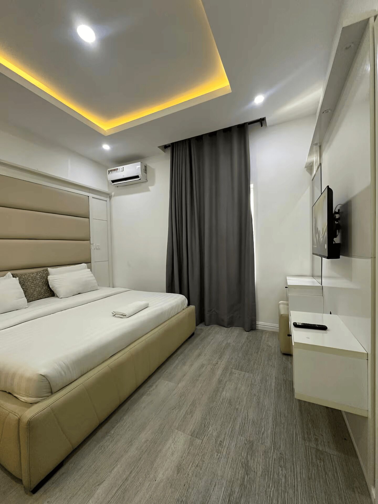 A modern hotel room with a large bed, a bedside table, a TV mounted on the wall, and an air conditioning unit.