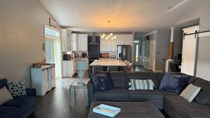 A modern living room with a grey sectional sofa, a glass coffee table, and a dining area with a kitchen in the background. - Spacious 4-bedroom, waterfront townhome at Lake Gaston (Henrico)