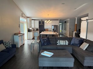 Ein modernes Wohnzimmer mit einem grauen Ecksofa, einem Glastisch und einem Essbereich, im Hintergrund ist eine Küche erkennbar.