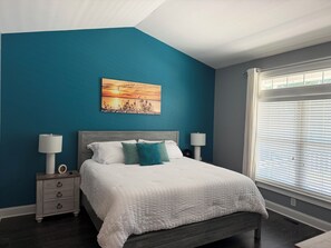Ein Schlafzimmer mit einem Bett, zwei Nachttischen, einem Wandbild und einem Fenster mit Jalousien.