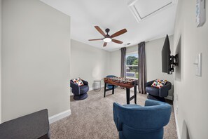 Ein Zimmer mit Deckenventilator, einem Kicker, zwei Sesseln und Blick ins Freie durch ein Fenster.