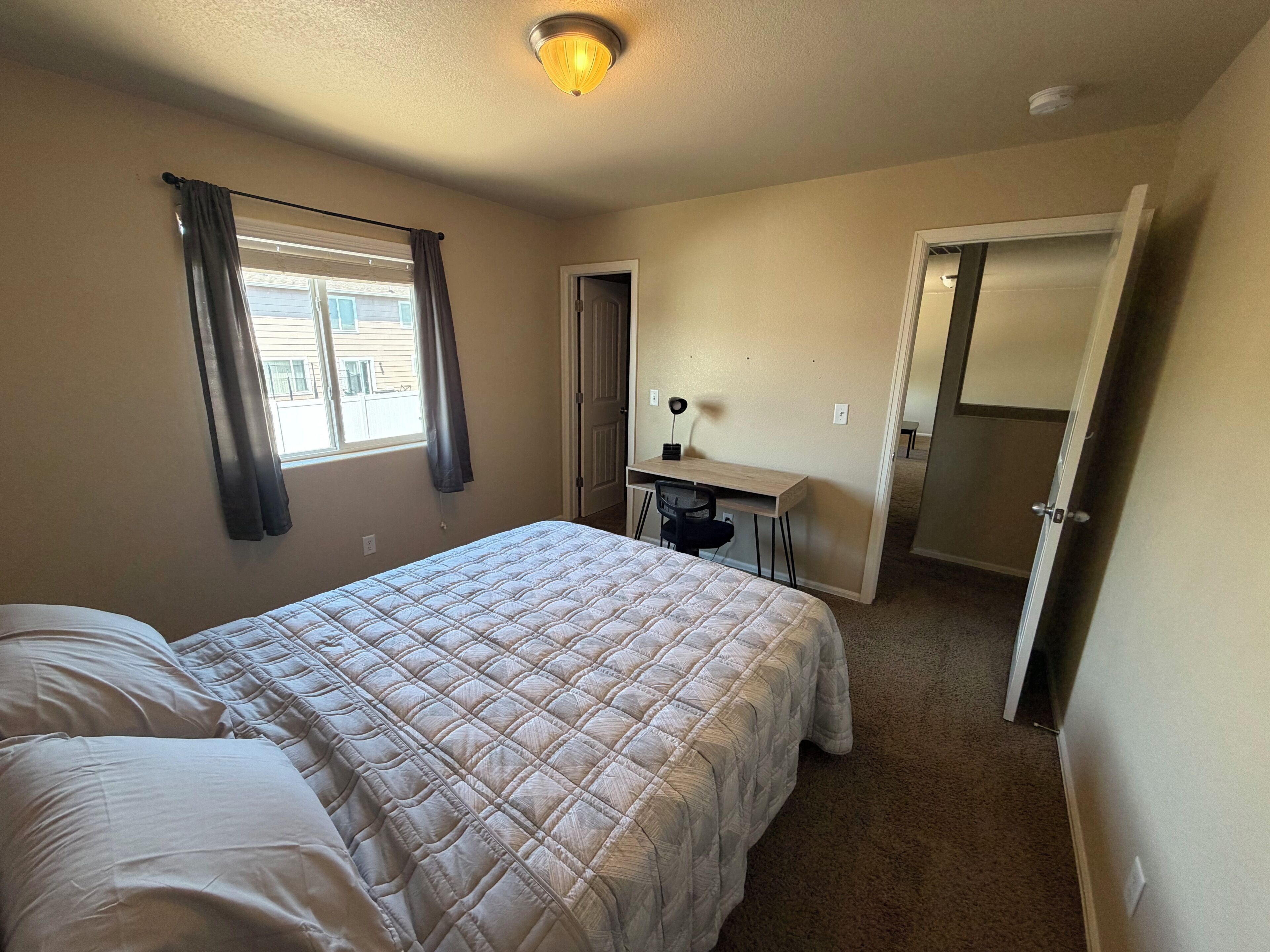 A bedroom with a bed, a desk, and a chair. There is a window with curtains, a door, and a ceiling light.