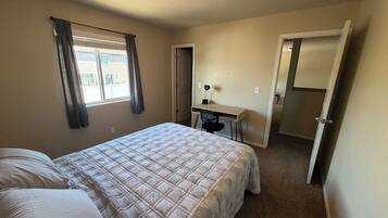 A bedroom with a bed, a desk, and a chair. There is a window with curtains, a door, and a ceiling light.