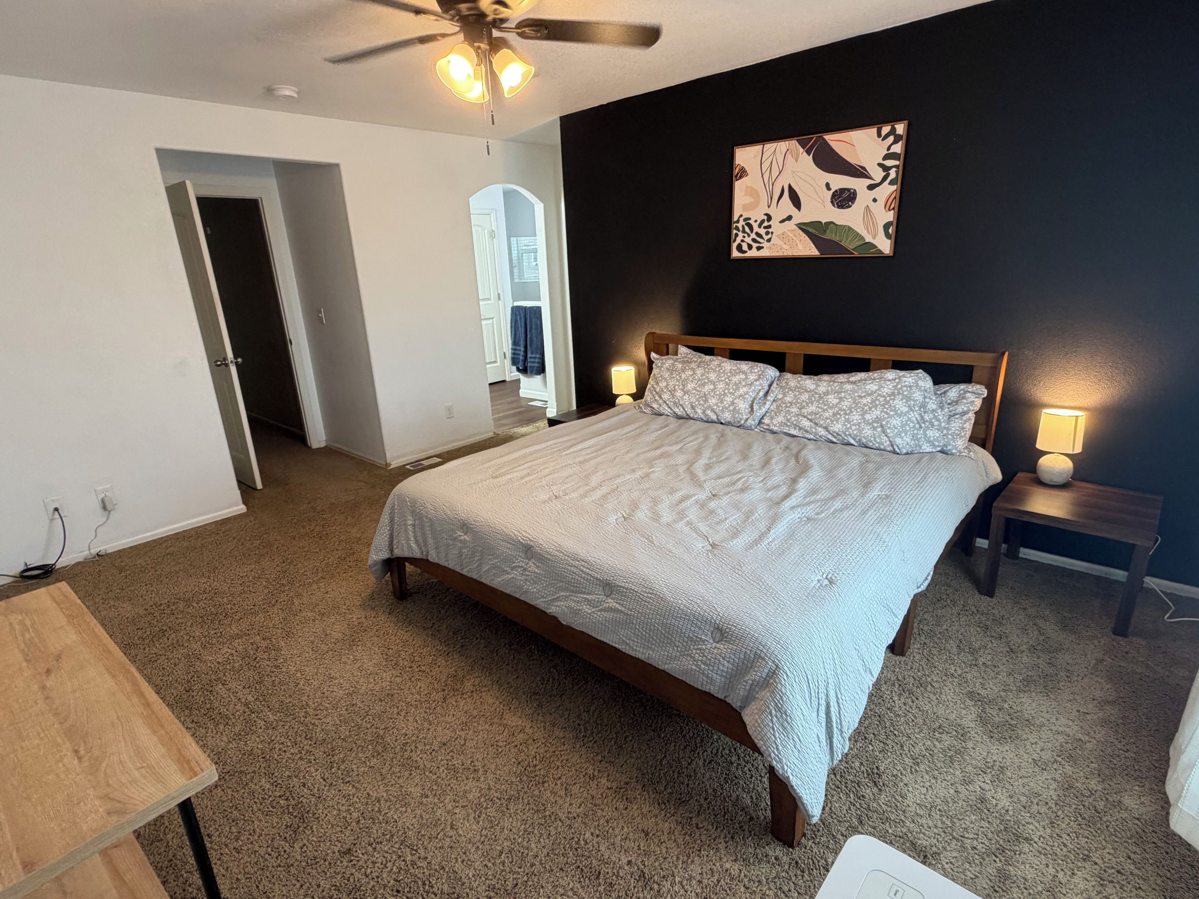 A bedroom with a bed, a ceiling fan, a nightstand, and a wall-mounted lamp.