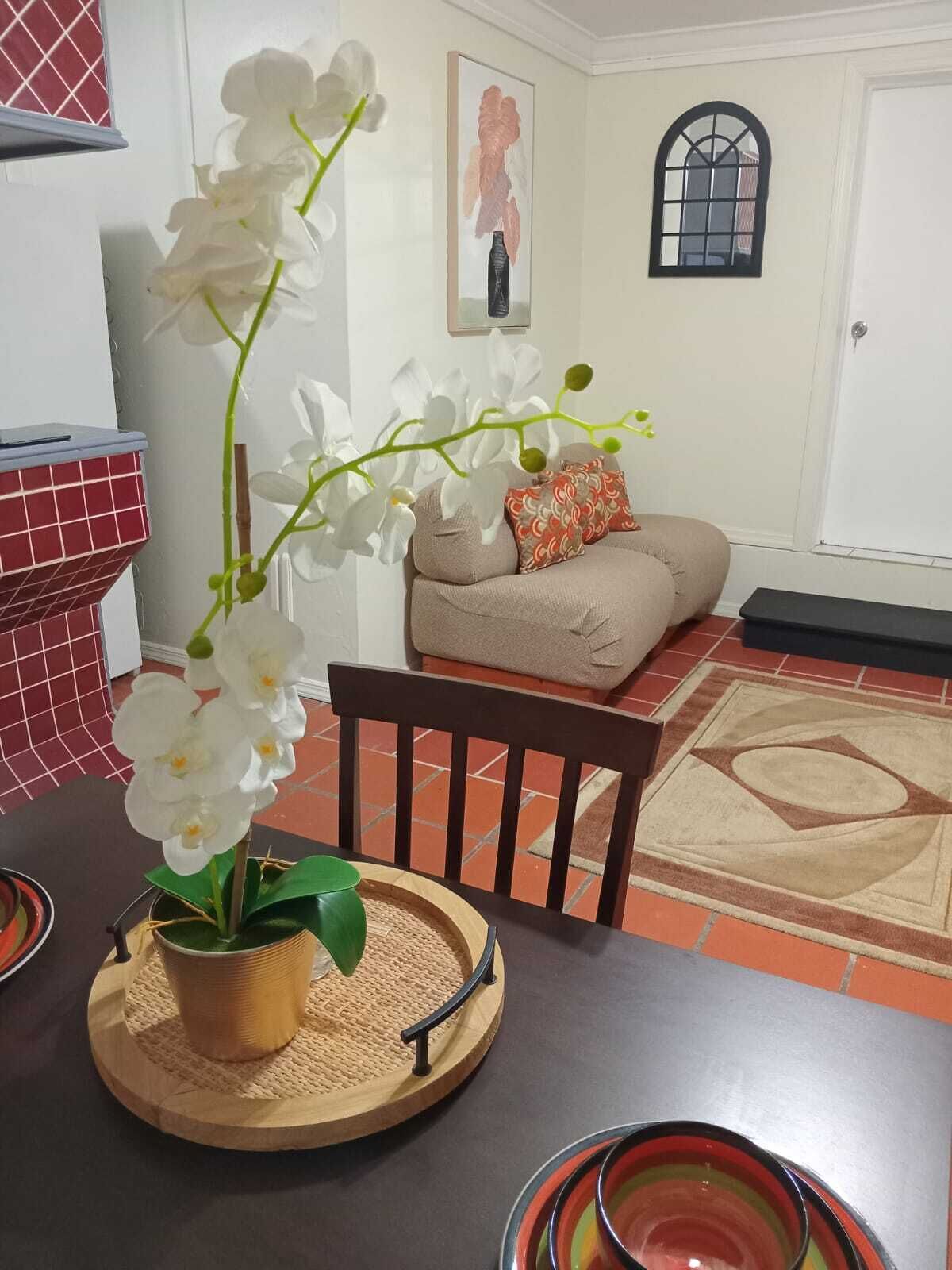 A dining area with a table set for two, a potted orchid, a wooden chair, a sofa, and a patterned rug.