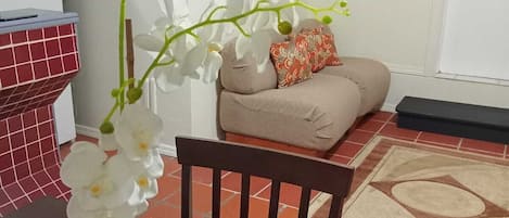 A dining area with a table set for two, a potted orchid, a wooden chair, a sofa, and a patterned rug.