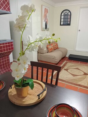 A dining area with a table set for two, a potted orchid, a wooden chair, a sofa, and a patterned rug. - Cozy 1-bdrm with ocean views, short walk to beach & shops! (Choc)