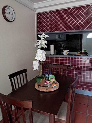A dining area with a wooden table, chairs, a clock on the wall, and a kitchen in the background. - Cozy 1-bdrm with ocean views, short walk to beach & shops! (Choc)