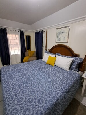 A bedroom with a large bed, a wooden headboard, a nightstand, a chair, and a window with curtains. - Cozy 1-bdrm with ocean views, short walk to beach & shops! (Choc)