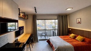 Une chambre d’hôtel comprenant un lit, un bureau, une chaise, une télévision, une fenêtre donnant sur des immeubles et un balcon.