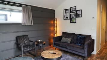 A modern living room with a leather sofa, a wooden coffee table, and a grey armchair. There are framed pictures on the wall and a window with curtains.