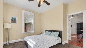 Quarto com cama, ventilador de teto, janela com persianas e porta para outro cômodo.