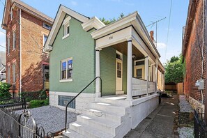Ein zweistöckiges Haus mit grüner Fassade, einer Veranda mit weißen Geländern und einer Ziegelaußenwand.