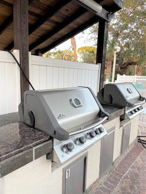 Ein Grill mit vier Brennern und einem digitalen Thermometer, vor einem weißen Zaun und Bäumen.