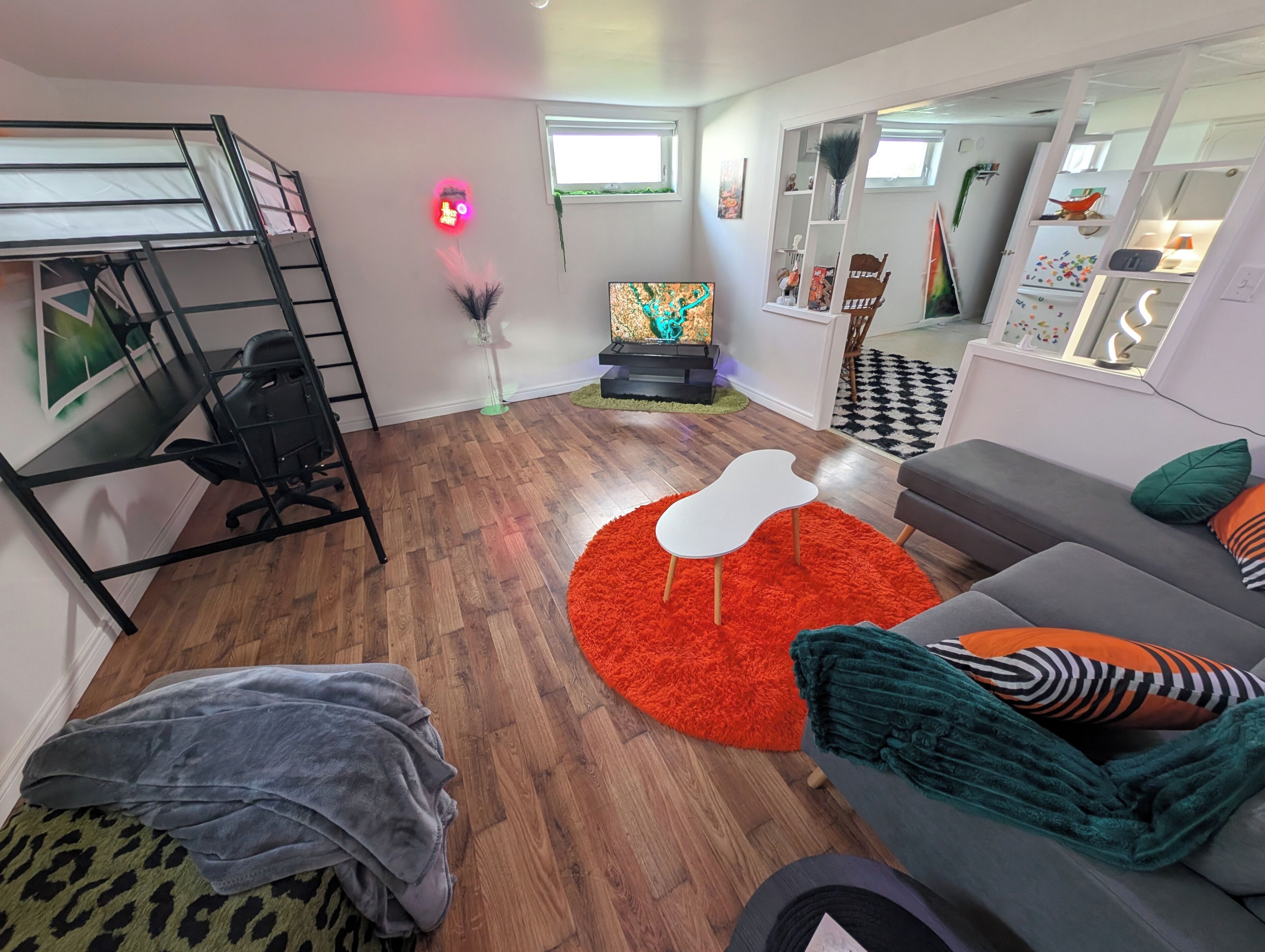 Un salon moderne avec un lit superposé, un canapé gris agrémenté de coussins colorés, un tapis rouge et une petite table blanche.