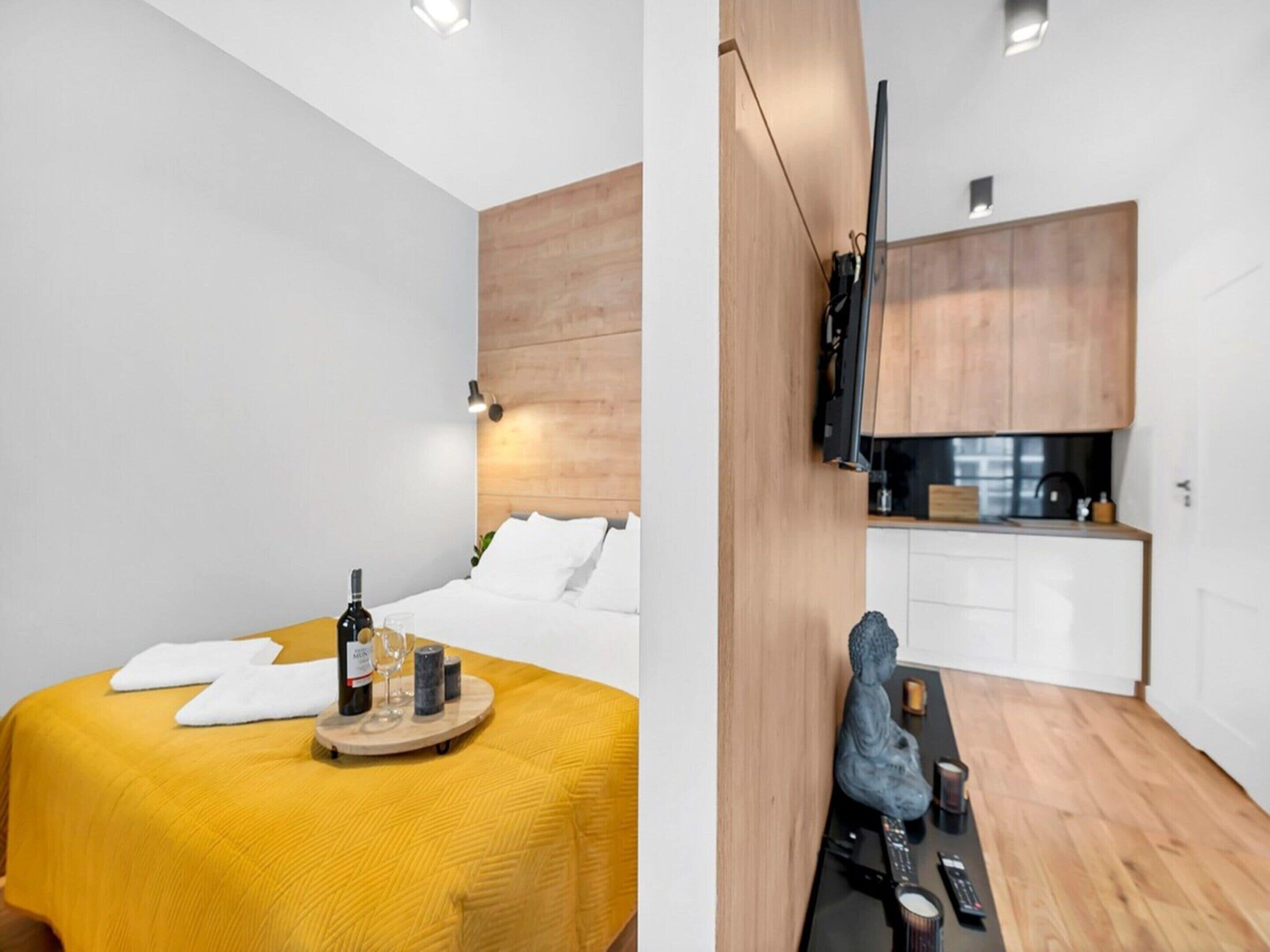 A modern hotel room with a bed, a small table with a bottle and glasses, a wooden headboard, and a kitchenette with cabinets and a sink.