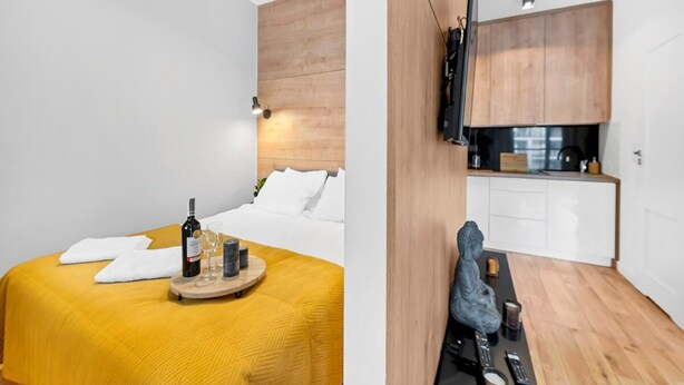 Ett modernt hotellrum med en säng, ett litet bord med en flaska och glas, en träpanel vid sänggaveln och ett litet kök med skåp och en diskho.