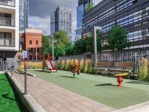 Ein Spielplatz mit Rutsche und Hydrant, umzäunt mit Bänken, mit Gebäuden im Hintergrund.