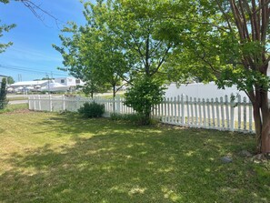 A grassy yard with a white picket fence, a tree, and a white building in the background. - Blue Anchor Beach House (Middle Township)