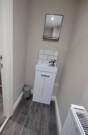 A narrow hallway with a white cabinet, a small trash bin, and a radiator. - Modern 2-Bed Ground Floor Flat | Sleeps 6 | Free Parking | Thetford (Norfolk)