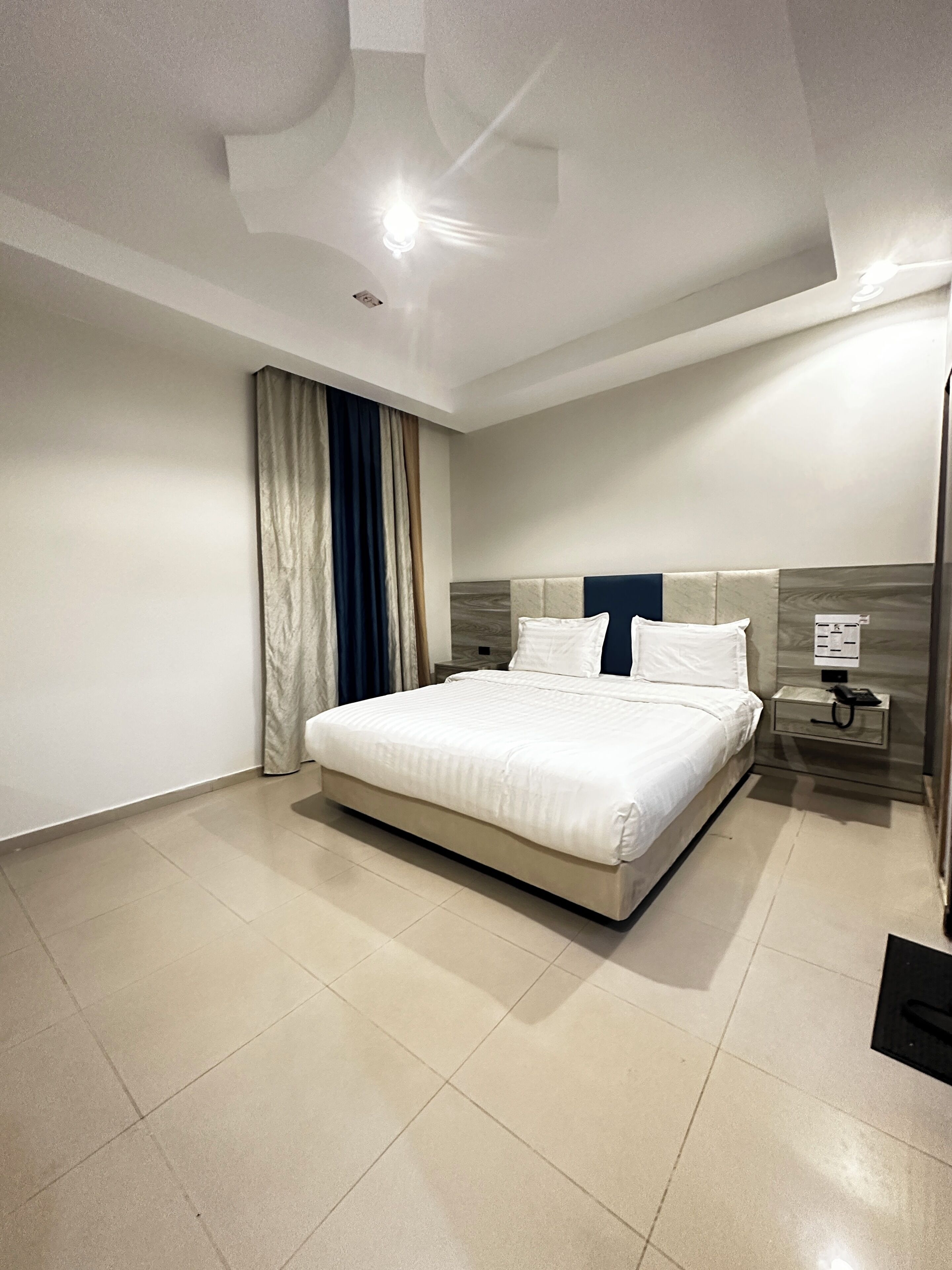 A modern hotel room with a large bed, a bedside table with a phone, and a wall-mounted light fixture.