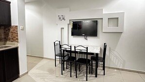 A modern living room with a dining area, a flat-screen TV mounted on the wall, and a kitchen area with dark cabinets.