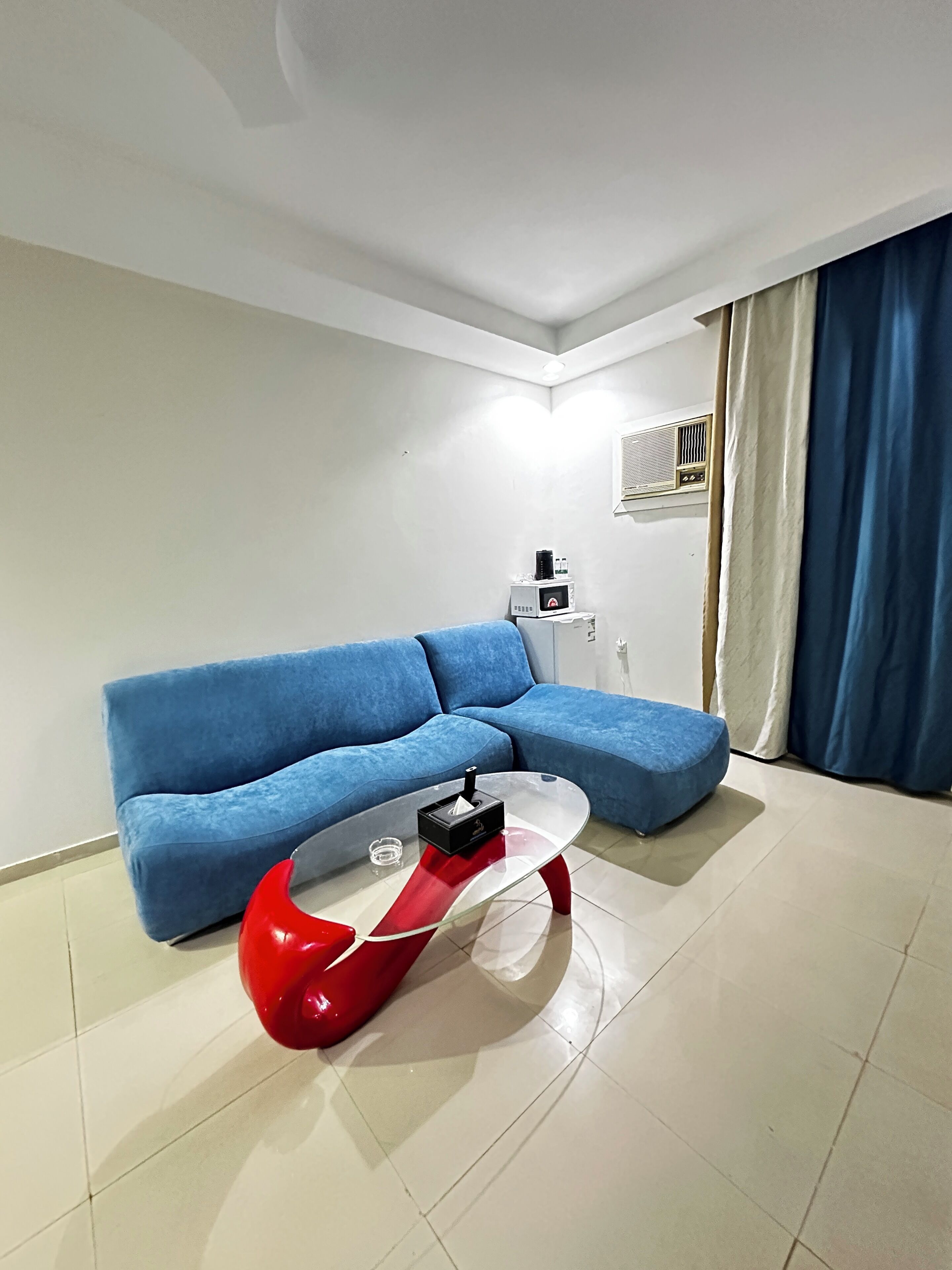 Ein Wohnzimmer mit einem blauen Sofa, einem roten und transparenten Couchtisch und einer weißen Klimaanlage an der Wand.