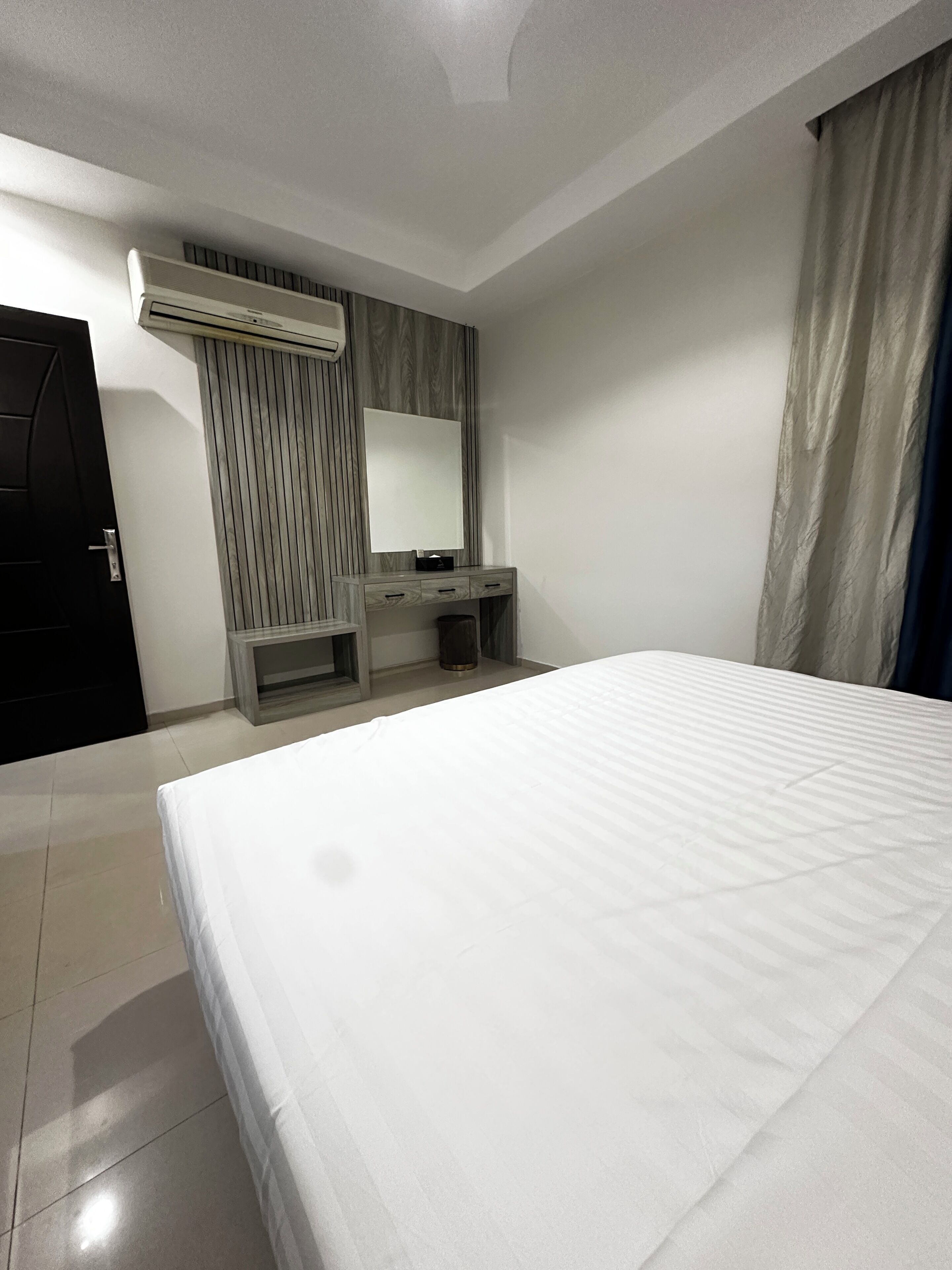 Quarto de hotel com uma cama grande, uma escrivaninha com cadeira, um espelho e um aparelho de ar condicionado.