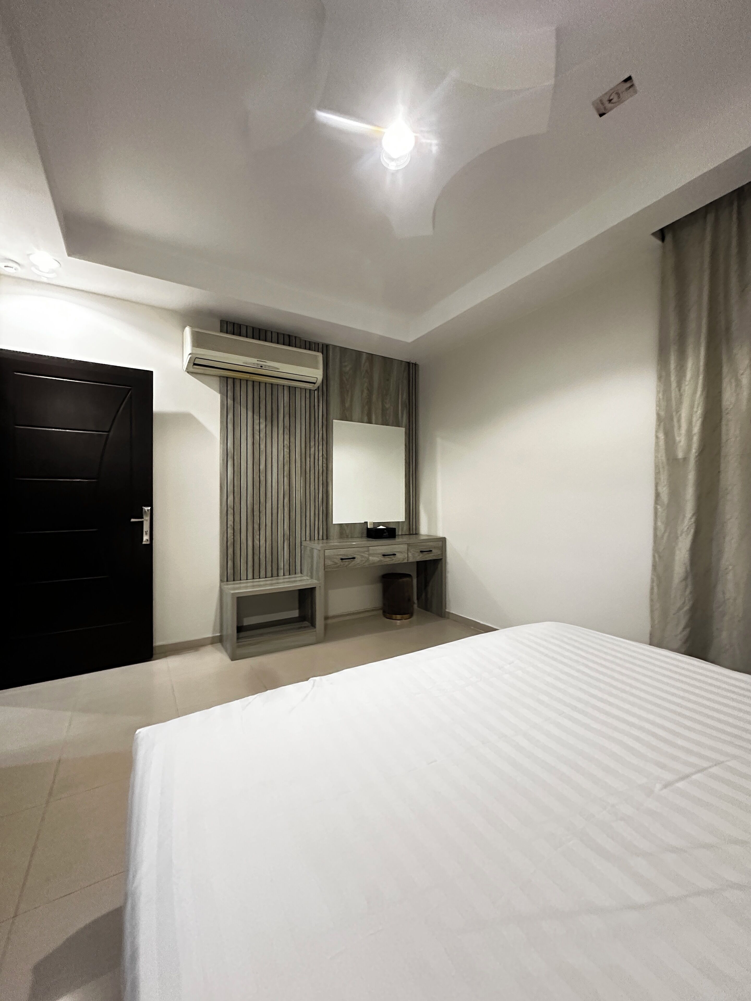 A hotel room with a bed, a desk with a mirror, an air conditioning unit, and a door.