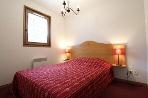 Ein Schlafzimmer mit einem Holz-Kopfteil, einer roten, gemusterten Bettdecke, zwei Nachttischlampen, einem Fenster mit Jalousien und einem an der Wand befestigten Heizkörper.