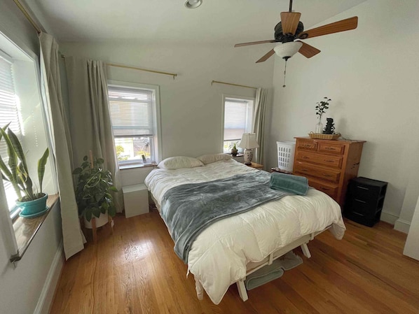 A bedroom with a bed, dresser, ceiling fan, windows, and potted plants.