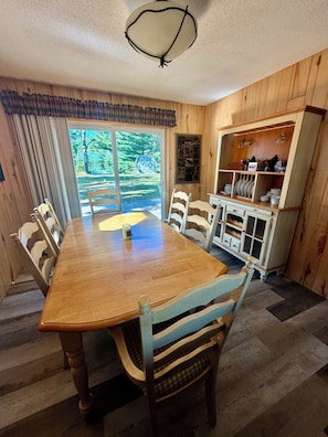 A dining area with a wooden table and chairs, a window with a view of trees, and a cabinet with dishes. - Lake Alexander Hideaway - Waterfront, Private Dock (Cushing)