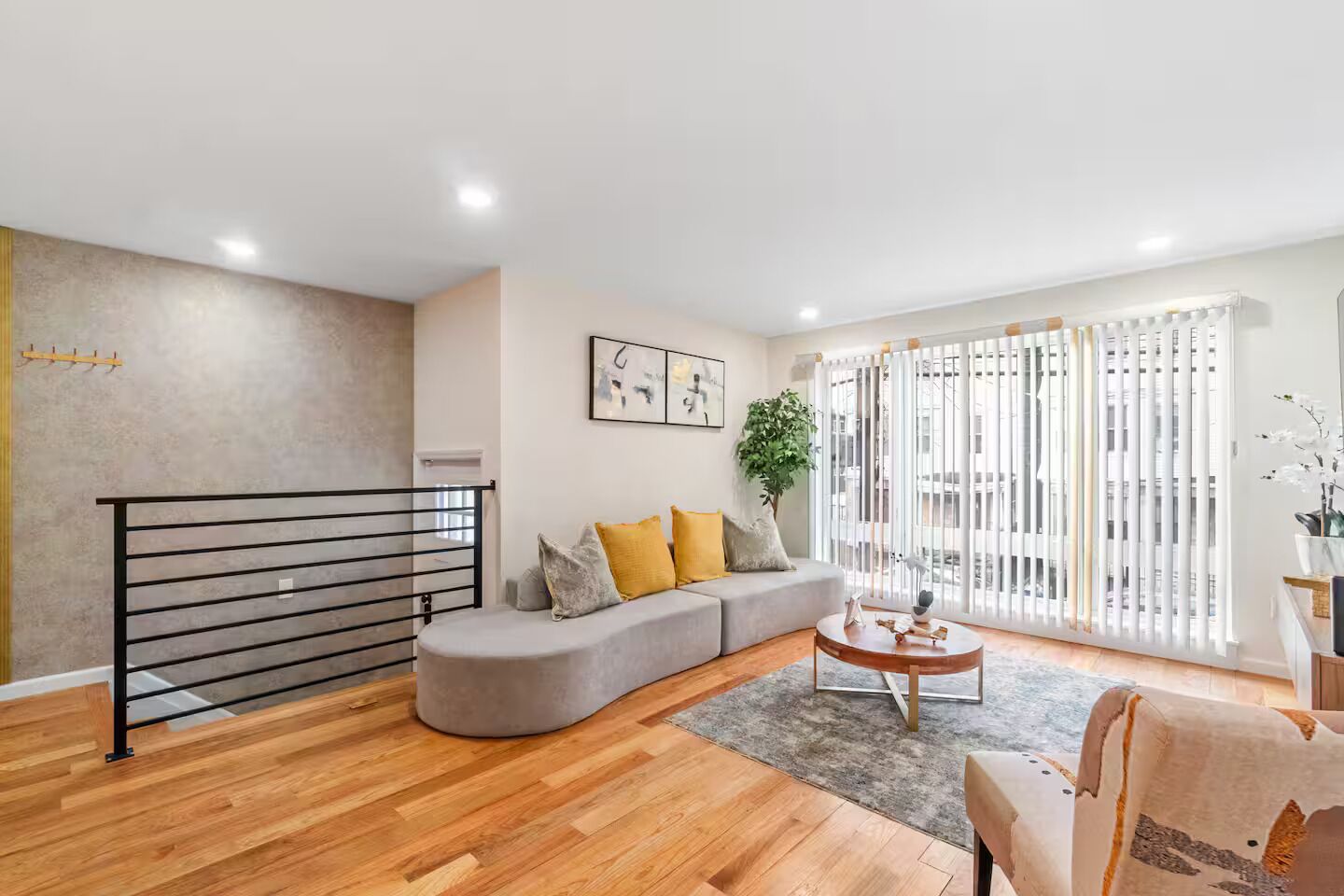 Un salón moderno con un sofá gris curvo, suelo de madera, barandilla de metal negro y un amplio ventanal con persianas.