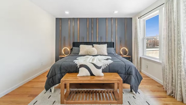 Un dormitorio moderno con una cama grande, lámparas de noche, un banco de madera y vistas a la ciudad a través de la ventana.