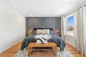 Um quarto moderno com uma cama grande, luminárias de cabeceira, um banco de madeira e vista da cidade pela janela.