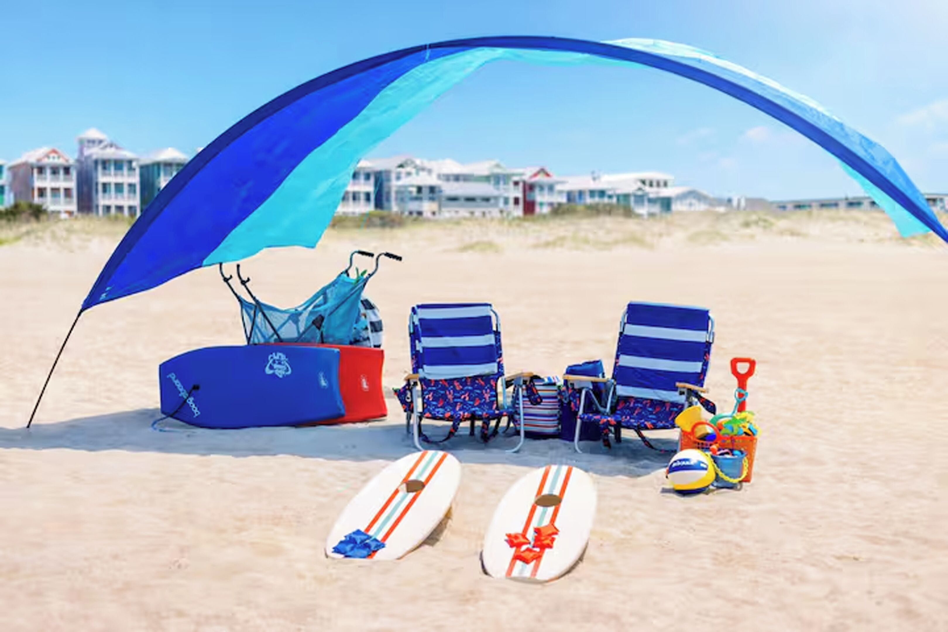 A beach scene with a blue canopy, two beach chairs, a surfboard, a paddleboard, and a beach ball.