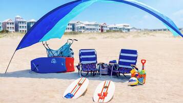 A beach scene with a blue canopy, two beach chairs, a surfboard, a paddleboard, and a beach ball.