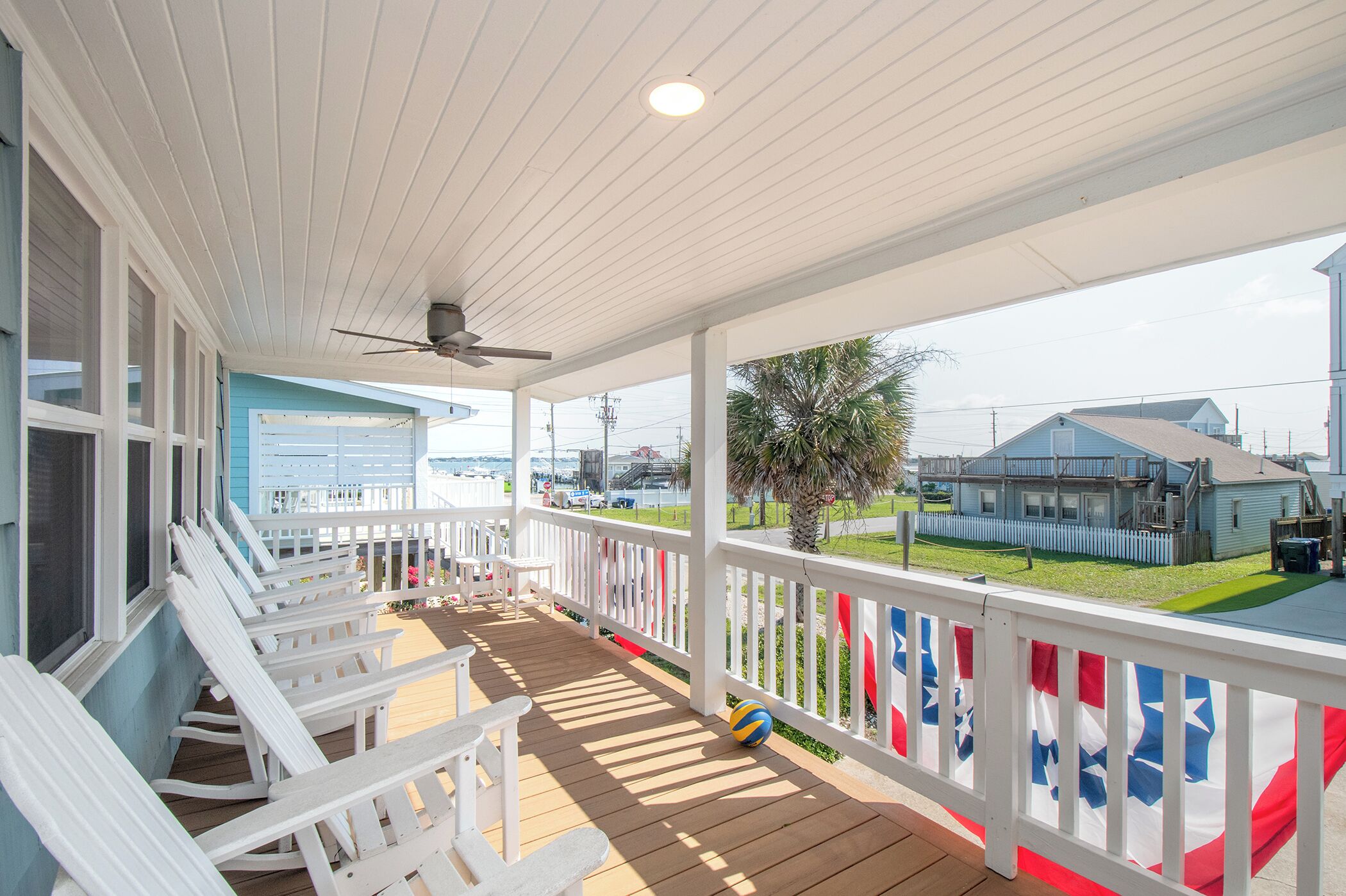 A covered porch with white railings and a ceiling fan, overlooking a residential area with houses and a clear sky.