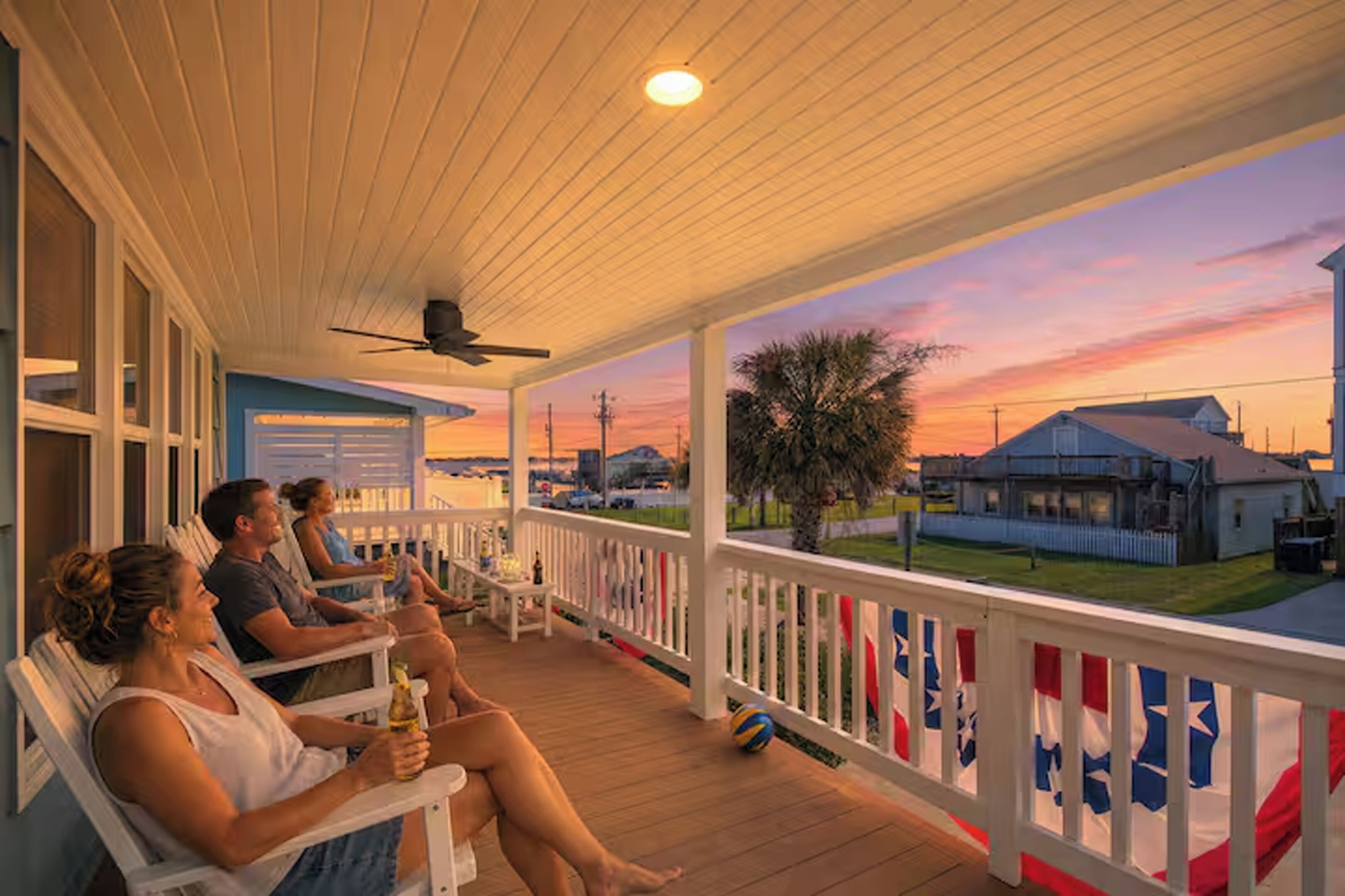 A covered porch with people seated, a ceiling fan, and a view of houses and a sunset.