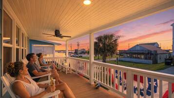 A covered porch with people seated, a ceiling fan, and a view of houses and a sunset.